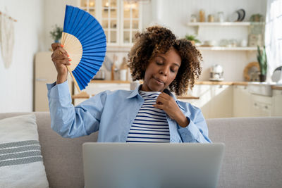 Exhausted overheated african american girl freelancer waving hand fan and working on laptop at home