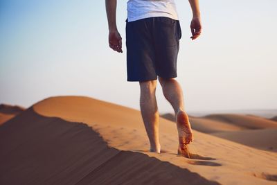 Low section of mid adult man walking on sand at desert against clear sky during sunset