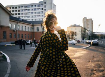 Portrait of young woman wearing sunglasses and overcoat on city street during sunset