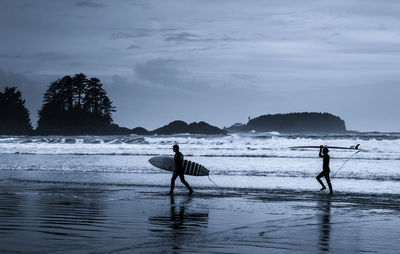 Silhouette people with surfboard at beach against sky