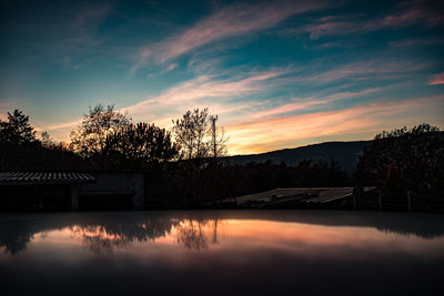 Silhouette houses by trees against sky during sunset