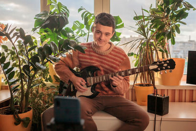 Young musician in cozy sweater sitting on table and playing electric guitar live in front of camera