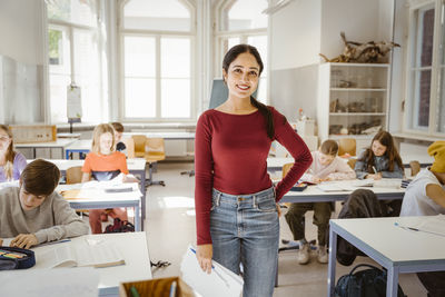 Portrait of smiling female teacher standing with hand on hip amidst students in classroom