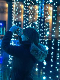 Side view of woman photographing by lights at night