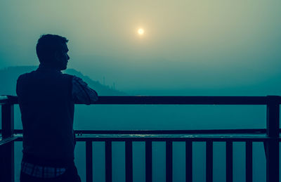 Rear view of man standing by railing against mountains during sunrise