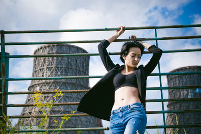 Low angle view of young woman standing by fence against silo