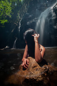 Rear view of woman sitting on rock against waterfall