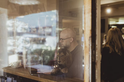 Bald man sitting with dog while using laptop seen through cafe window