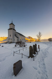 Snow covered wooden posts on field by building against sky during sunset