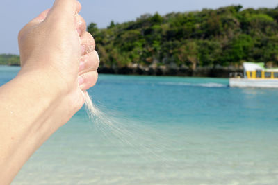 Cropped image of hand holding sand over sea against mountain