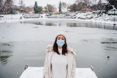 Relaxed woman with eyes closed wearing face mask standing on pier in front of frozen lake in city