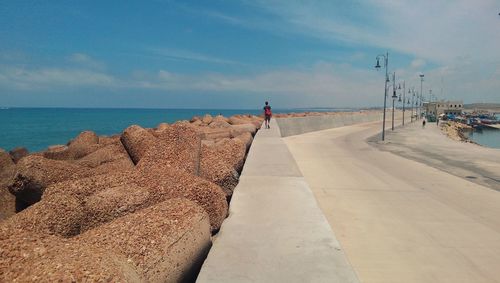 Rear view of man walking on retaining wall by tetrapods at sea shore against sky