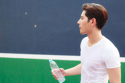 Young man looking away while holding plastic bottle against wall