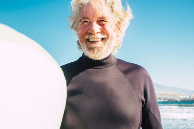Smiling senior man standing with surfboard at beach on sunny day