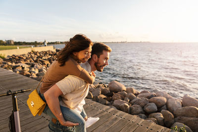 Man piggybacking happy girlfriend while enjoying on promenade by sea during vacation