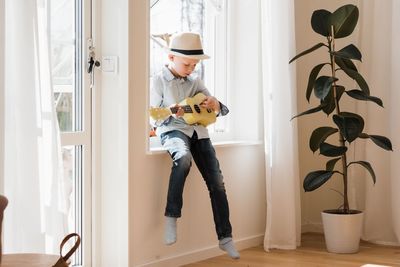 Young boy sat on a window ledge at home playing ukulele