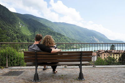 Rear view of couple sitting on bench looking at mountains against sky