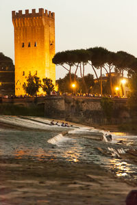 Illuminated building by sea against sky at sunset