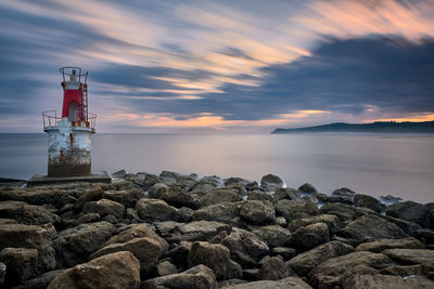 Lighthouse on rocks by sea against sky during sunset