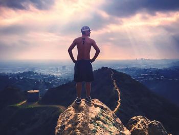 Rear view of shirtless man standing on mountain with hand on hip against cloudy sky