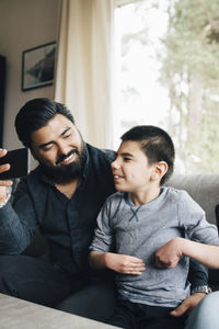 Smiling father holding mobile phone looking at autistic son while sitting on sofa in living room