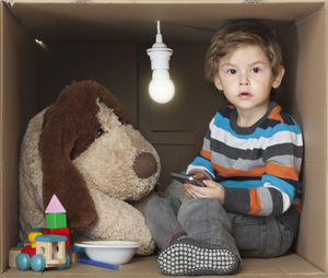 Boy holding mobile phone while sitting in cardboard box against white background