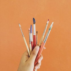 Cropped image of hand holding various paintbrushes against orange background