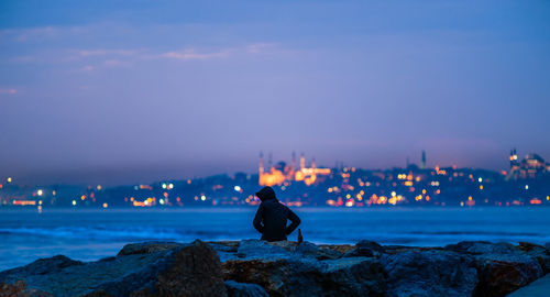 Hooded person sitting at rocky shore against sky at dusk