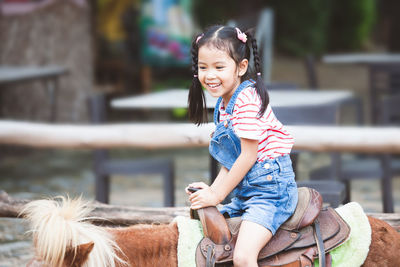 Side view of cute cheerful girl sitting on pony at farm