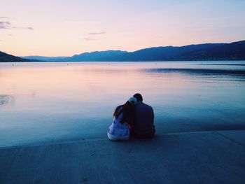 Rear view of couple sitting on shore against sky during sunset