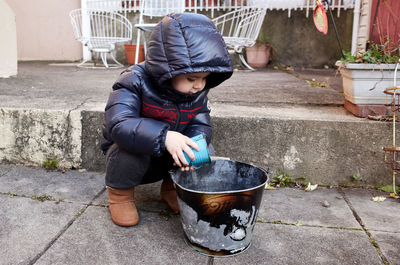 Curious toddler boy in a winter suit playing with a bucket in the backyard