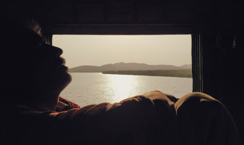 Man relaxing by lake against sky seen through window