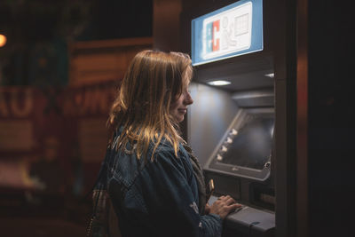 Side view of smiling young woman using atm at night