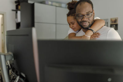 Smiling freelancer working on computer with daughter hugging him from behind at home