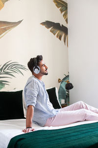 Side view of thoughtful bearded hispanic male in headphones sitting on bed and enjoying good music at home