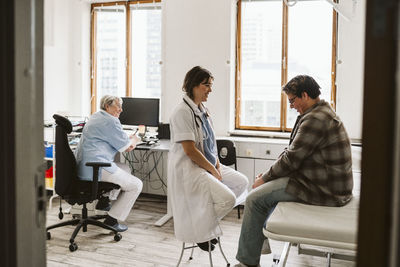 Smiling female healthcare worker talking with young male patient while senior nurse sitting at desk