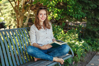 Portrait of happy young woman reading book while sitting on bench at park
