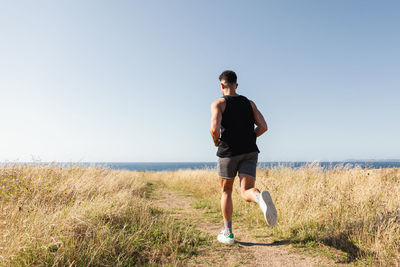 Muscular male jogger running along path in meadow during training on background of sea in summer