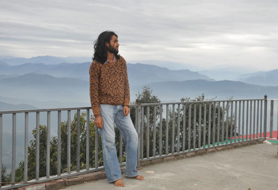 A indian young man with long hair looking sideways while standing against mountains in background