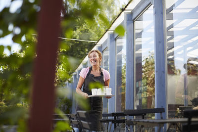 Happy senior waitress carrying disposable glasses on tray at sidewalk cafe