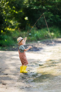 Preschooler boy in yellow rubber boots plays fishing, child fishes with stick lake in countryside