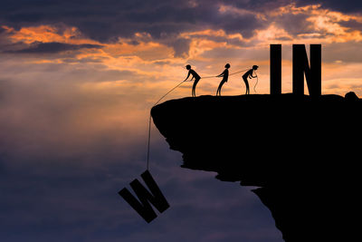 Low angle view of silhouette figurines with alphabets on cliff against cloudy sky during sunset