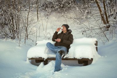Full length of man drinking coffee while sitting on snow covered bench