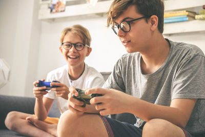 Smiling brothers holding remote control while sitting on sofa at home