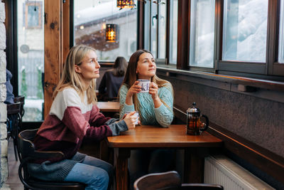 Two women in winter sweaters drink hot tea in a bar and look out the window at snowy weather.