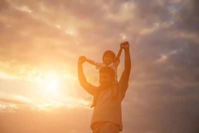 Low angle view of man piggybacking daughter while standing against sky during sunset