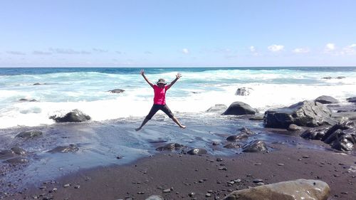 Cheerful young woman with arms raised jumping at beach against sky