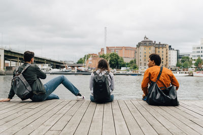 Rear view of friends sitting on pier by river in city against sky