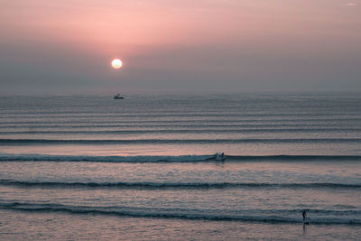 Scenic view of sea with surfers against sky during sunset