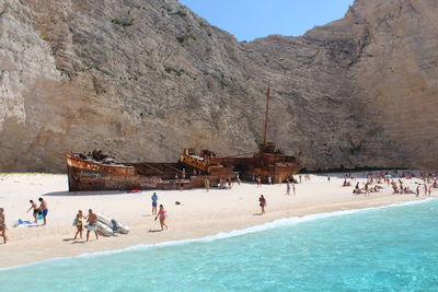 People at beach by shipwreck against rock formation